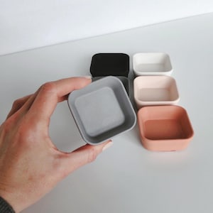 May include: Six small square ceramic dishes in various colours, including black, white, grey, pink, and orange. The dishes are arranged in a row on a white surface.
