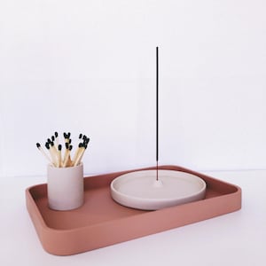 Op de afbeelding: Een terracotta-kleurige rechthoekige tray bevat een kleine cilindrische container met lucifers en een witte keramische wierookhouder. Een wierookstokje staat rechtop in de houder. De items staan op een wit oppervlak.