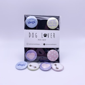 May include: A set of five dog-themed pins. The pins are round and feature different designs, including a heart with the word "DOGS" inside, a paw print pattern, and the text "Life is better with dogs". The pins are packaged in a clear plastic bag with a white card that says "DOG LOVER PIN SET" and includes the social media handles "@giannarosedesigns_" and "@giannarosedesigns".