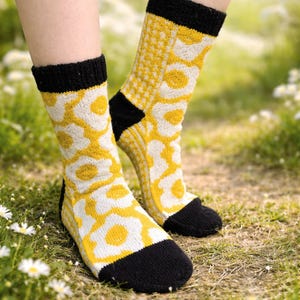 Peut inclure: Paire de chaussettes tricotées avec un motif floral. Les chaussettes présentent un motif de fleurs jaunes et blanches sur fond crème, avec des poignets, des talons et des orteils noirs. Les chaussettes sont portées à l'extérieur sur de l'herbe.