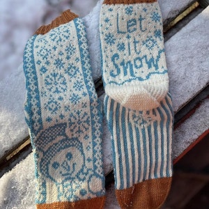 Könnte beinhalten: Ein Paar gestrickte Wintersocken mit einem Schneemann-Design und dem Schriftzug "Let it Snow". Die Socken haben ein blau-weißes Schneeflockenmuster, eine Schneemann-Grafik und braune Bündchen und Zehen. Die Socken liegen auf einer schneebedeckten Oberfläche.