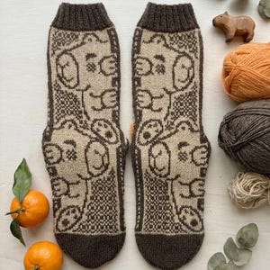 May include: A pair of mid-calf knit socks featuring a repeating pattern of cartoon bear faces in dark brown and beige. The socks have a ribbed cuff and a solid dark brown toe section, showcasing a detailed jacquard knit design throughout the fabric.
