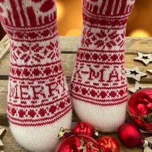 Op de afbeelding: Paar rood-witte gebreide sokken met een kerstpatroon. De sokken liggen op een houten oppervlak met rode en gouden ornamenten.