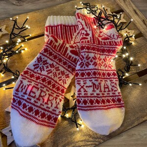 Op de afbeelding: Een paar witte en rode gebreide sokken met een kerstpatroon. De sokken liggen op een houten oppervlak met kerstverlichting.