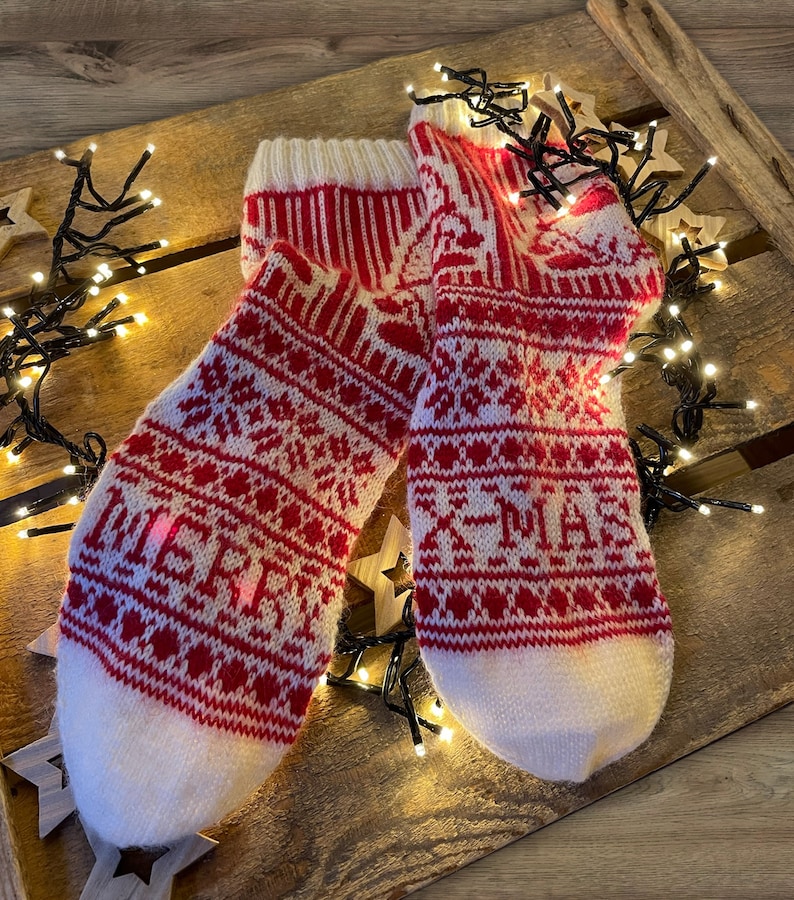 Op de afbeelding: Een paar witte en rode gebreide sokken met een kerstpatroon. De sokken liggen op een houten oppervlak met kerstverlichting.