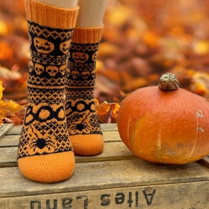 Op de afbeelding: Oranje en zwarte gebreide sokken met een Halloween-thema met pompoenen en geesten. De sokken liggen op een houten oppervlak met een pompoen op de achtergrond.
