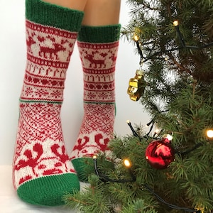 May include: A pair of red, white, and green Christmas socks with a reindeer pattern. The socks are worn by a person standing near a decorated Christmas tree.