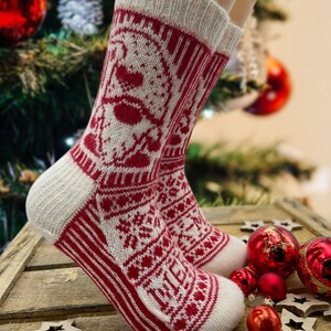 Op de afbeelding: Witte en rode gebreide sokken met een kerstthema. De sokken hebben een sneeuwvlok, een rendier en een kerstboom.