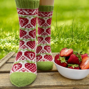 Könnte beinhalten: Ein Paar grüne, weiße und rote gestrickte Socken mit einem Erdbeermuster. Die Socken liegen auf einer Holzkiste mit einer Schüssel Erdbeeren davor.