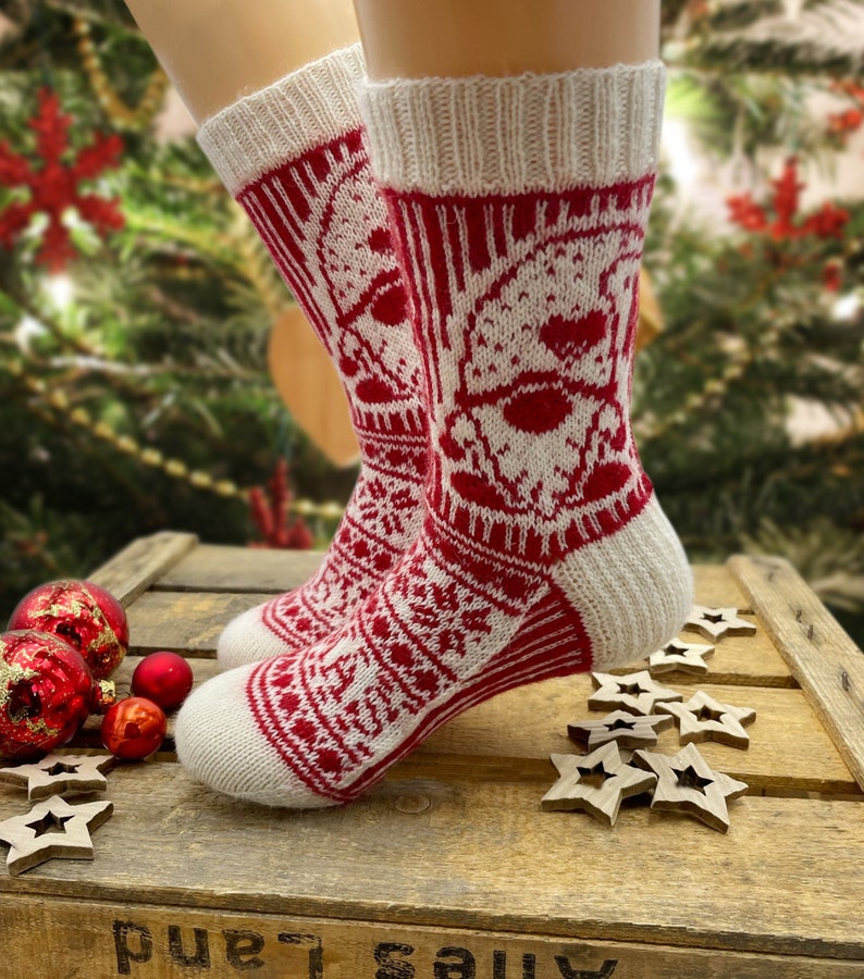 Op de afbeelding: Een paar witte en rode gebreide sokken met een kerstthema. De sokken hebben een wit sneeuwvlokkenpatroon en een rood kerstmangezicht.