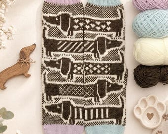 Puede incluir: Un par de calcetines de punto con un patrón repetitivo de perros salchicha de color marrón oscuro sobre un fondo crema. Un calcetín tiene el puño y la puntera de color lavanda, mientras que el otro es de color azul claro. Los perros tienen patrones.