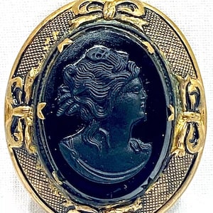 May include: An oval cameo brooch with a black profile of a woman in a gold-toned frame. The frame has a textured border and bow details, creating a vintage aesthetic. The brooch is a classic piece of jewellery.