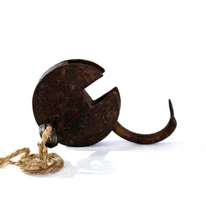 May include: A rusty, antique metal hook with a circular body and a curved hook. A small metal piece and a length of rope are attached. The hook is likely for hanging or securing items. The overall color is brown.