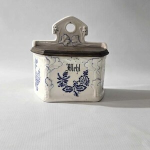 May include: A white ceramic wall-mounted container with a dark brown lid. The container is decorated with blue floral patterns and the word "Mehl" in a decorative font. The top has a decorative design with a hole.
