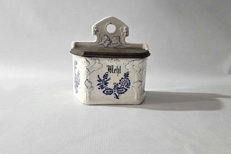 May include: A white ceramic wall-mounted container with a dark brown lid. The container is decorated with blue floral patterns and the word "Mehl" in a decorative font. The top has a decorative design with a hole.