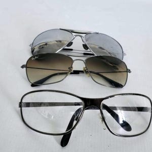 Puede incluir: Se muestran tres pares de gafas sobre un fondo blanco. El par superior son gafas de sol de aviador plateadas. El par del medio son gafas de sol degradadas marrones. El par inferior son gafas con montura negra y lentes transparentes.