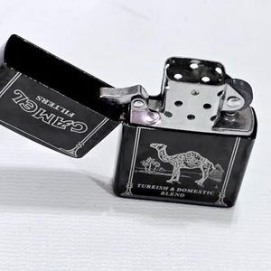 Może przedstawiać: Czarna metalowa zapalniczka z napisem "CAMEL" i "FILTERS" wydrukowanym na pokrywie. Korpus zapalniczki przedstawia grafikę wielbłąda z napisem "TURKISH & DOMESTIC BLEND". Zapalniczka jest otwarta, odsłaniając srebrny mechanizm zapłonu.