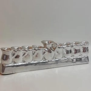 Puede incluir: Una menorá plateada con nueve candelabros en forma de pequeñas jarras. La menorá se encuentra sobre una base rectangular y se exhibe sobre una superficie blanca.