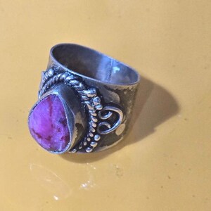 Può includere: Un anello d'argento con una grande gemma magenta a forma di goccia. L'anello presenta un design ornato con dettagli intrecciati e perlati attorno alla gemma e alla fascia. L'anello è posto su uno sfondo giallo.