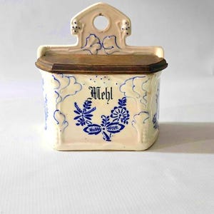 May include: Cream-colored ceramic container with a wooden lid, featuring blue floral designs and the word "Mehl." The container has a decorative top with a hole for hanging. A vintage kitchen storage item.