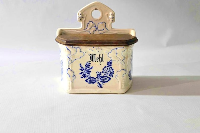 May include: Cream-colored ceramic container with a wooden lid, featuring blue floral designs and the word "Mehl." The container has a decorative top with a hole for hanging. A vintage kitchen storage item.