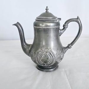 May include: An antique silver-coloured teapot with a curved spout and handle. The teapot has a lid and is decorated with an ornate floral design on the body. The teapot is likely made of metal.