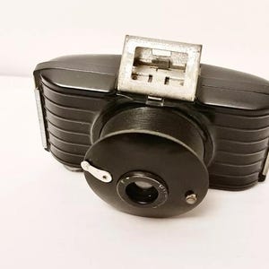 May include: A vintage black camera with a silver viewfinder. The camera has a ribbed body and a circular lens. The lens is covered by a black cap. The camera is likely from the mid-20th century.