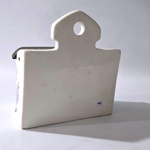 May include: A white ceramic wall pocket with a house-shaped top and a rectangular body. The pocket has a small hole for hanging and a dark-colored rim. The design is simple and minimalist, suitable for small plants or decorative items.