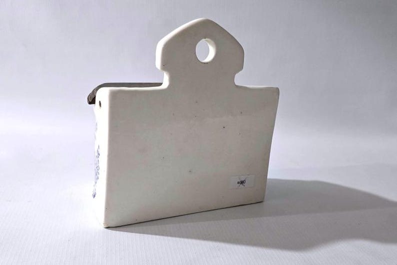 May include: A white ceramic wall pocket with a house-shaped top and a rectangular body. The pocket has a small hole for hanging and a dark-colored rim. The design is simple and minimalist, suitable for small plants or decorative items.