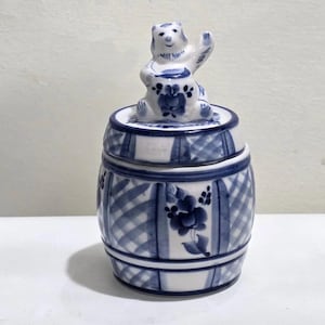 Op de afbeelding: Een keramische pot met deksel, met een blauw-wit ruitpatroon en bloemaccenten. Een klein berenfiguur zit bovenop het deksel en houdt een honingpot vast. De pot is tonvormig, met een traditioneel, handgeschilderd ontwerp.