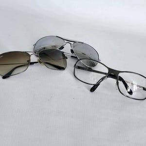 Puede incluir: Tres pares de gafas y gafas de sol se muestran sobre una superficie blanca. Las gafas de sol tienen montura plateada y lentes espejadas. Otro par tiene montura negra y lentes degradadas. El tercer par tiene montura negra y lentes transparentes.