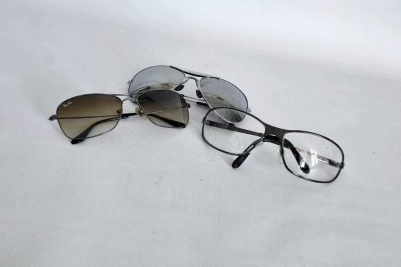 Puede incluir: Tres pares de gafas y gafas de sol se muestran sobre una superficie blanca. Las gafas de sol tienen montura plateada y lentes espejadas. Otro par tiene montura negra y lentes degradadas. El tercer par tiene montura negra y lentes transparentes.