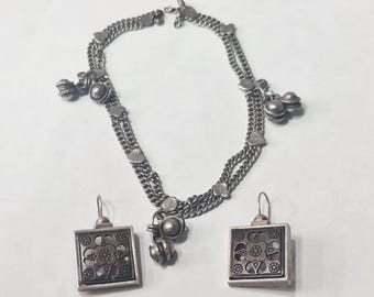 May include: A silver-toned jewelry set featuring a necklace and matching earrings. The necklace has multiple chain strands with heart-shaped and floral accents, and dangling bell charms. The square earrings have a floral design.