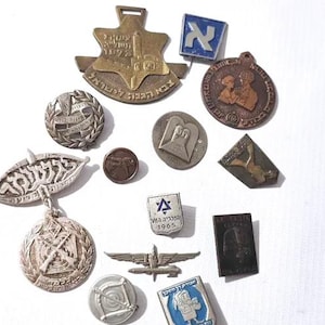 May include: A selection of vintage metal pins and medallions in various shapes and designs. Some feature Hebrew text, a Star of David, and other symbols. The pins are in shades of silver, bronze, and blue.