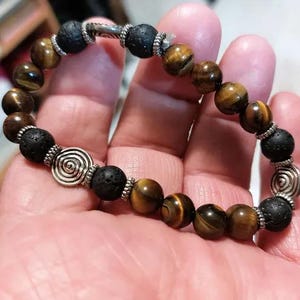 Pode incluir: Uma pulseira de contas com pedras de olho de tigre e lava negra alternadas. A pulseira inclui detalhes metálicos prateados com desenhos em espiral. A pulseira é segurada em uma mão.
