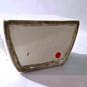 May include: A white ceramic vase with blue floral designs. The vase has a rectangular shape with a slightly angled base. The rim and base edges have a dark, aged appearance. A red dot and blue mark are visible on the base.