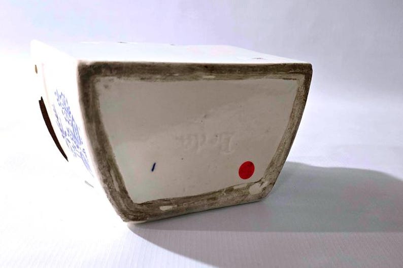 May include: A white ceramic vase with blue floral designs. The vase has a rectangular shape with a slightly angled base. The rim and base edges have a dark, aged appearance. A red dot and blue mark are visible on the base.