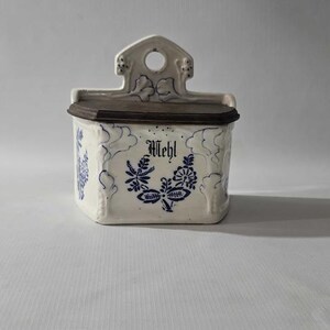 May include: A white ceramic container with a dark brown lid and a decorative top. The container is adorned with blue floral patterns and the word "Mehl" in black script. A vintage kitchen storage item.