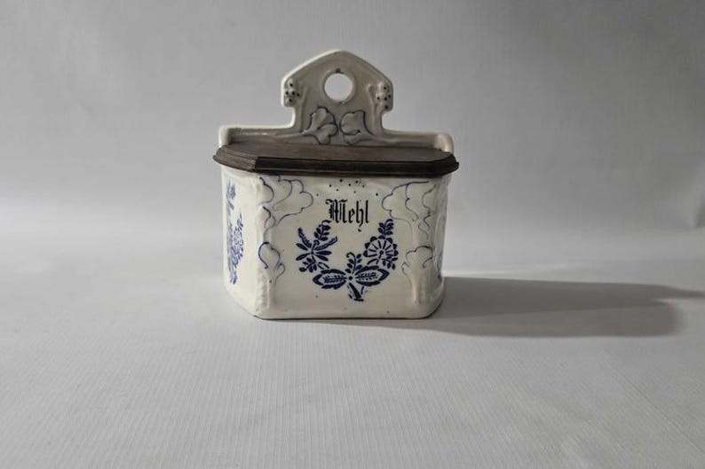 May include: A white ceramic container with a dark brown lid and a decorative top. The container is adorned with blue floral patterns and the word "Mehl" in black script. A vintage kitchen storage item.