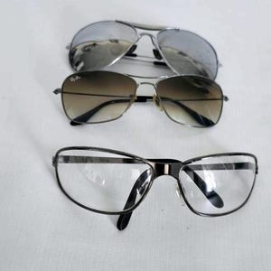 Puede incluir: Se muestran tres pares de gafas sobre un fondo blanco. El par superior presenta lentes plateadas espejadas, el par del medio tiene lentes degradadas marrones y el par inferior tiene lentes transparentes con montura negra.