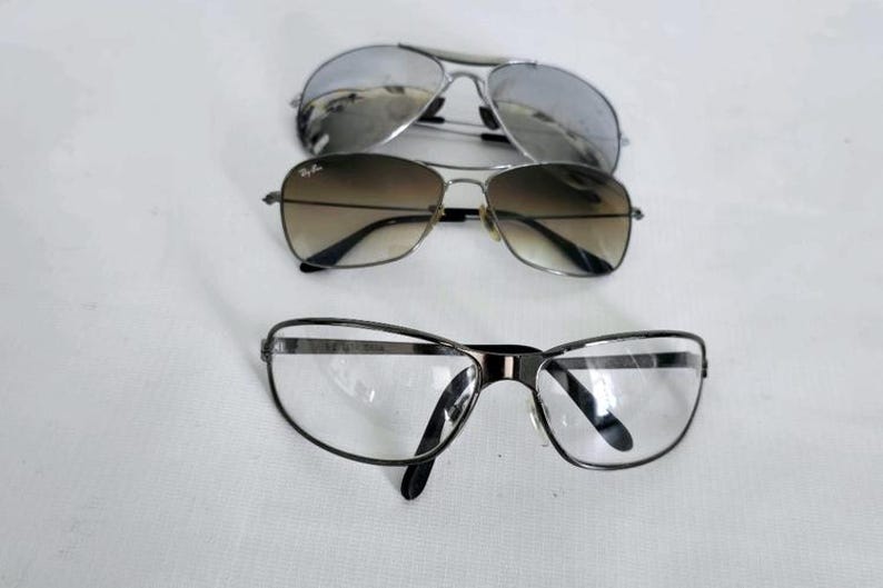 Puede incluir: Se muestran tres pares de gafas sobre un fondo blanco. El par superior presenta lentes plateadas espejadas, el par del medio tiene lentes degradadas marrones y el par inferior tiene lentes transparentes con montura negra.
