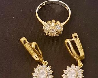 Könnte beinhalten: Ein goldfarbenes Schmuckset mit einem Ring und Ohrringen. Der Ring und die Ohrringe haben einen zentralen ovalen Stein, der von kleineren, baguetteförmigen Steinen umgeben ist, wodurch ein Sonnenstrahl-Design entsteht. Das Set wird auf dunklem Untergrund präsentiert.