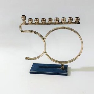 以下が含まれることがあります： 9つのキャンドルホルダーが付いたシルバー色のメノラー。濃い青色の長方形の台座に置かれています。デザインには2つの大きな円が組み込まれています。メノラーは、ユダヤ教のハヌカの祝日に使用される宗教的なアイテムです。