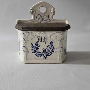 May include: A white ceramic wall-mounted container with a dark brown lid. The container is decorated with blue floral patterns and the word "Mehl" in black script. The top has a decorative handle with a hole.