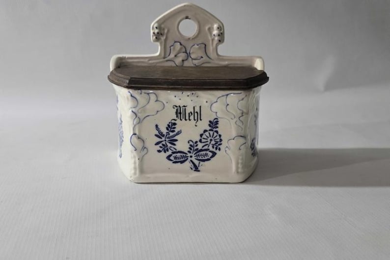 May include: A white ceramic wall-mounted container with a dark brown lid. The container is decorated with blue floral patterns and the word "Mehl" in black script. The top has a decorative handle with a hole.