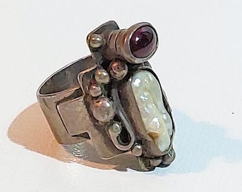 May include: A silver-toned ring with a unique design. It features a large, irregular pearl, a round, dark red gemstone, and several small, round accents. The ring has a geometric band and an overall artistic aesthetic.