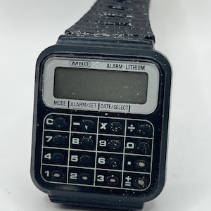 Puede incluir: Reloj calculadora negro con una pantalla digital y una banda negra. La esfera del reloj dice "MBO ALARM LITHIUM" y tiene botones para "MODE ALARM/SET DATE/SELECT". La calculadora tiene botones para "C", "-", "X", "÷", "7", "8", "9", "0", "4", "5", "6", "-", "1", "2", "3", "+", y "=".