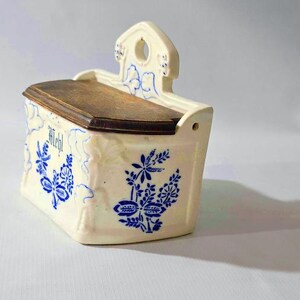 May include: Cream-colored ceramic wall-mounted container with a wooden lid. The container is decorated with blue floral patterns and the word "Mehl". It has a small hole for hanging.