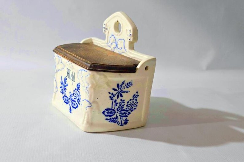 May include: Cream-colored ceramic wall-mounted container with a wooden lid. The container is decorated with blue floral patterns and the word "Mehl". It has a small hole for hanging.