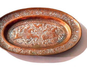 May include: Oval copper-colored tray with an embossed design. The tray features a central scene of figures on horseback and a detailed border. The copper has a slightly aged appearance, with some areas showing a lighter patina.